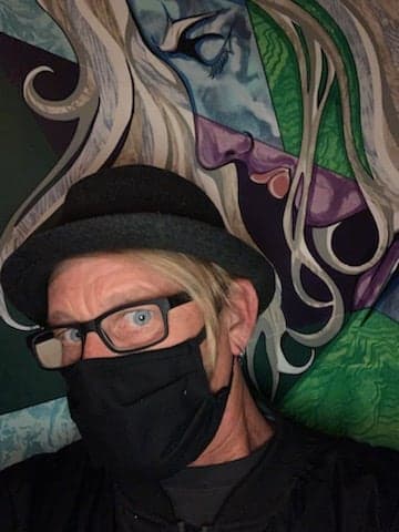 Sean Amundson in a hat, glasses and a black mask, standing in front of one of his paintings.
